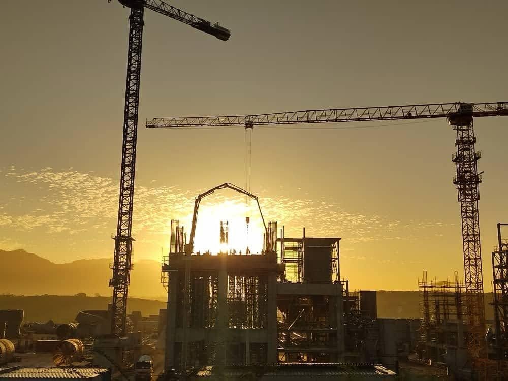 Dos grúas torre sobre una planta industrial en construcción, recortadas contra el sol del atardecer.