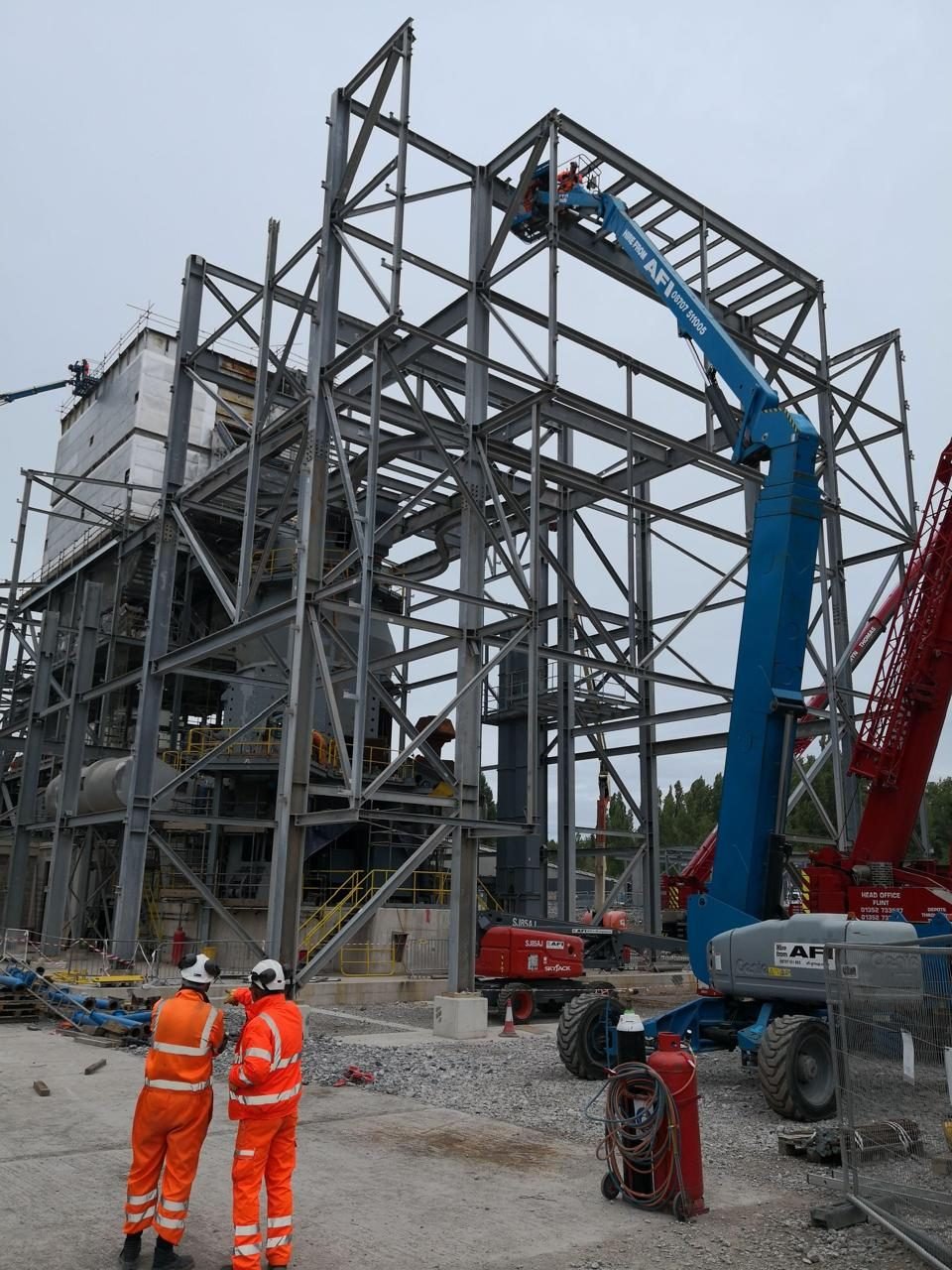 Dos técnicos con overol reflectivo y casco observan el montaje de una estructura metálica intervenida con plataforma elevadora.