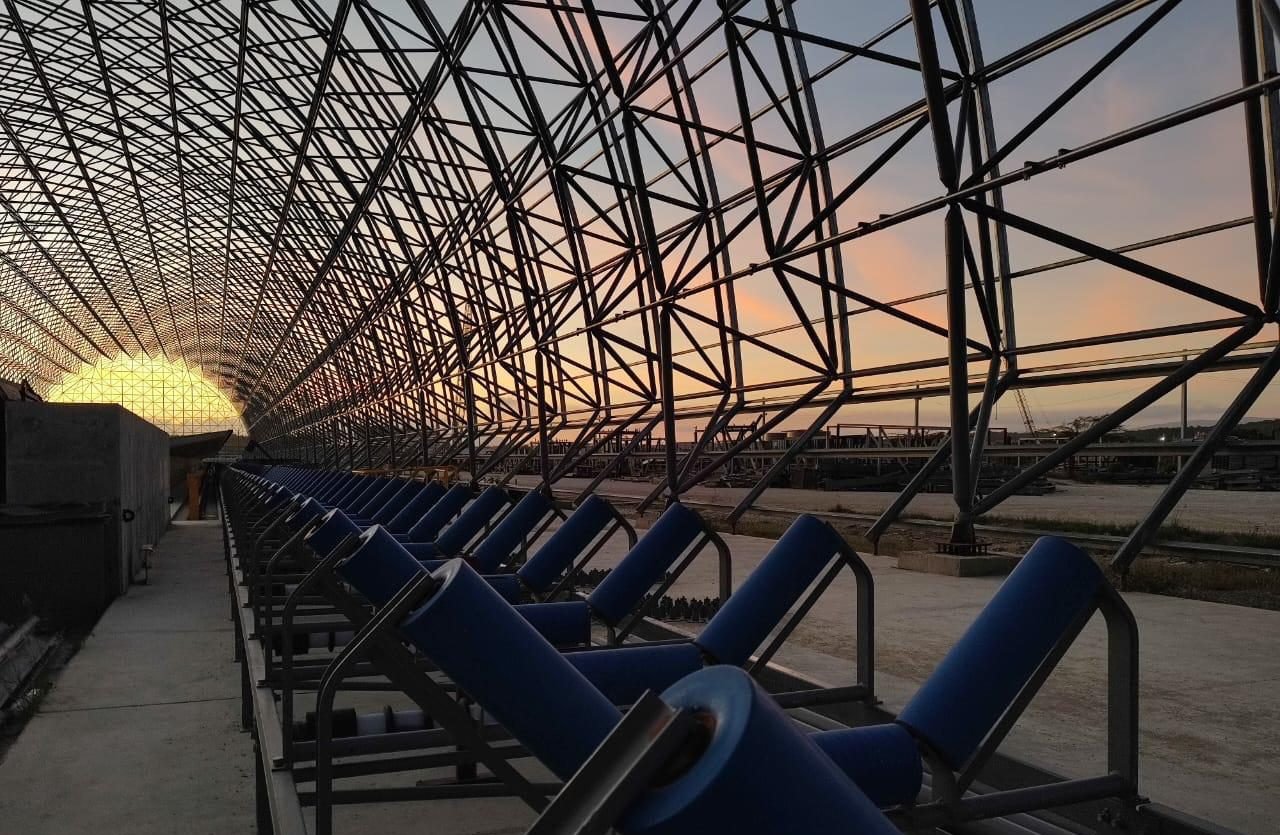 Línea de rodillos de una banda transportadora instalada bajo la estructura reticular del domo, al atardecer.